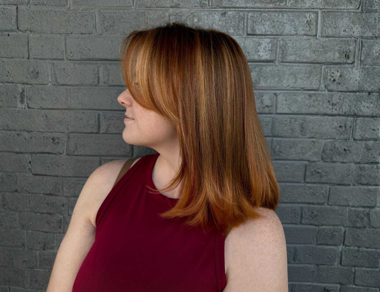 Red-haired woman with side bangs, wearing a maroon top, against a gray brick wall.